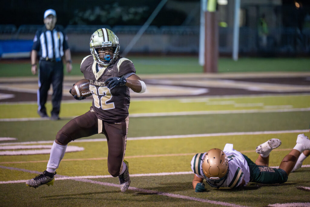 Football player number 12 in brown gold running with ball past fallen opponent in white green uniform on field.