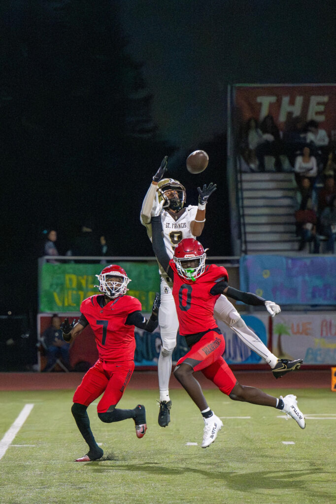 St. Francis football player in white jumps to catch ball as two defenders in red attempt to block.