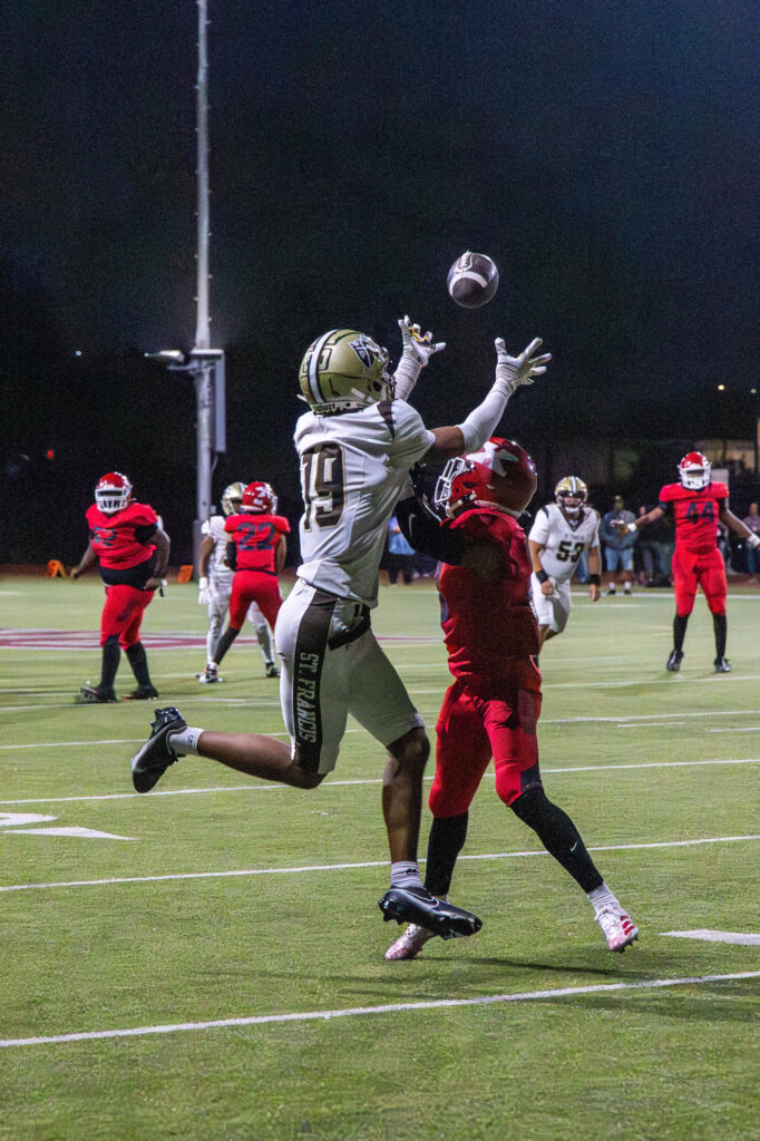 Two football players jump to catch the ball during a nighttime game on the field.