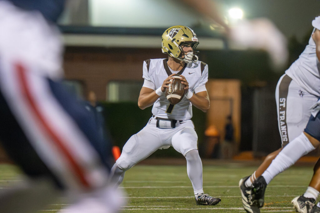 Football quarterback in white uniform and gold helmet prepares to throw ball during night game.