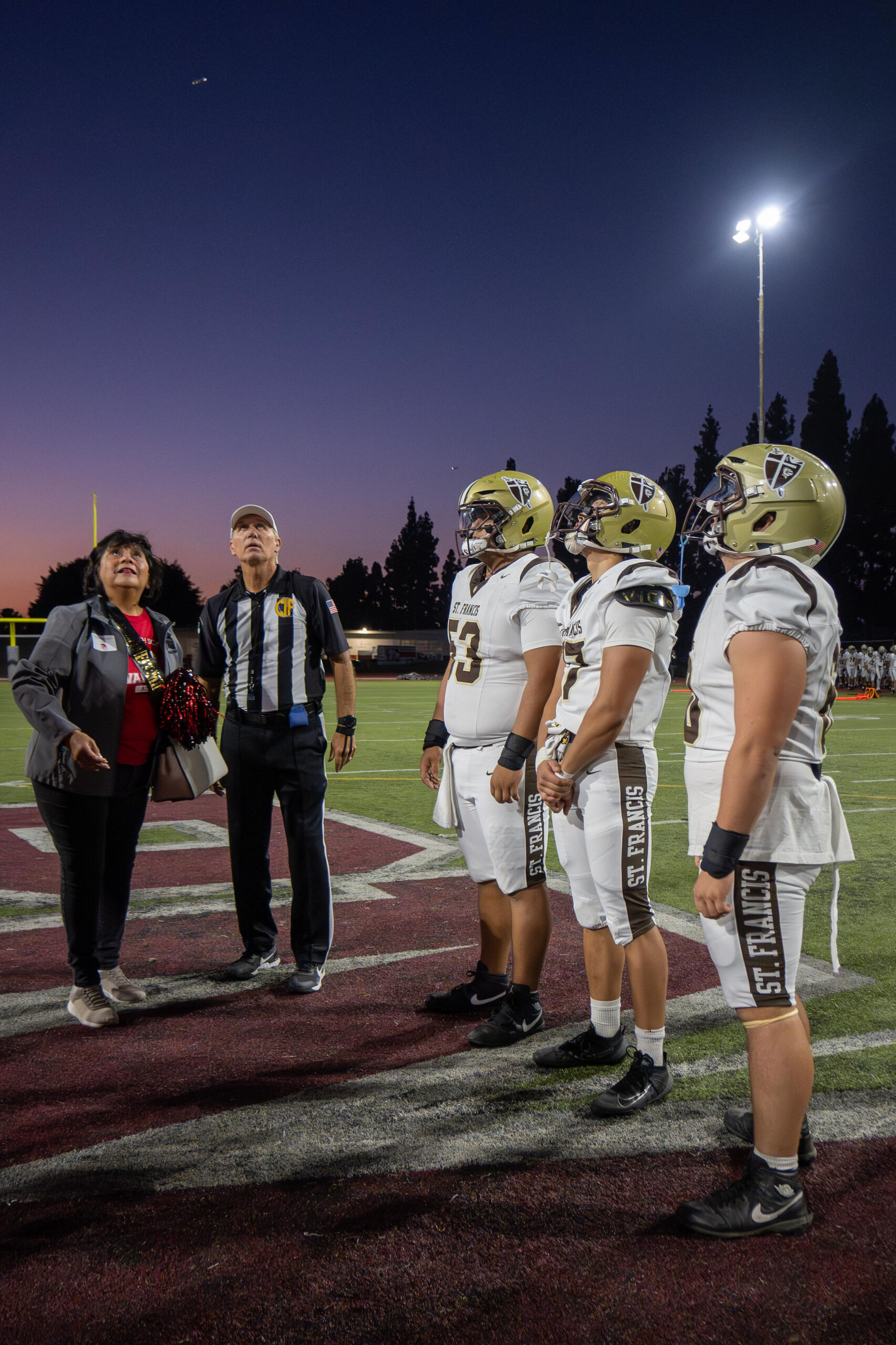 Three football players in white and gold uniforms with a referee and woman holding pom-poms at sunset.